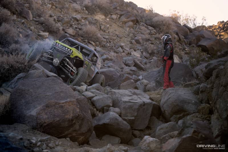 Vaughn Gittin Jr. winching with his co-driver out of the car by the rock that his winch line is tied to