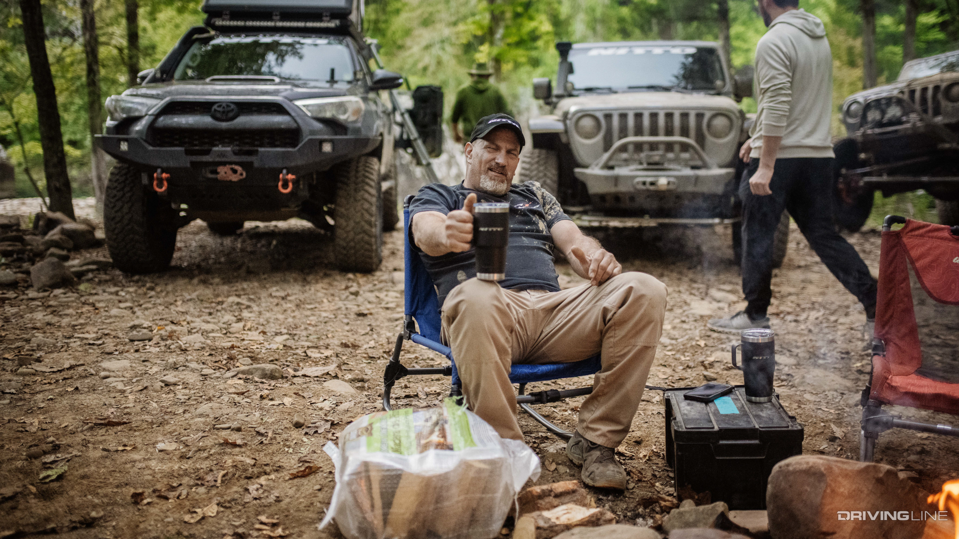 Man holding Nitto branded mug in front of off-road trucks in forest
