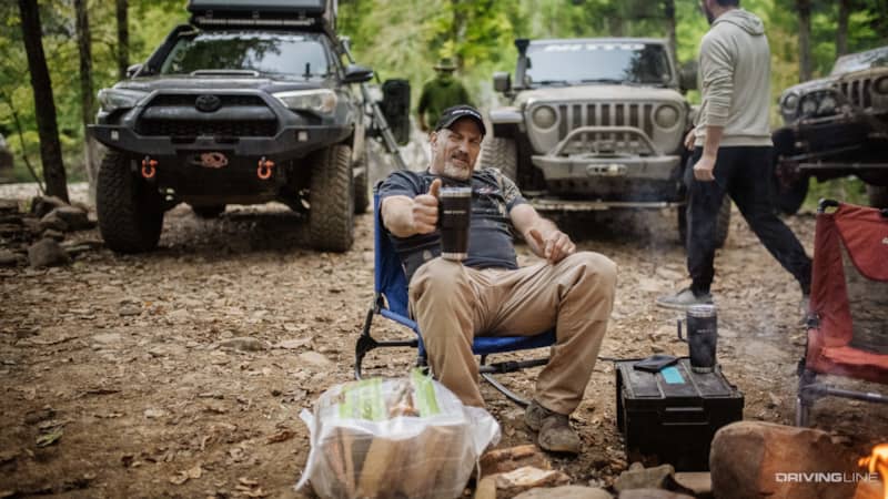 Man holding Nitto branded mug in front of off-road trucks in forest